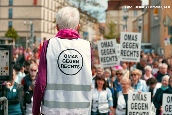 Eine Frau mit weißer Weste, Aufschrift „Omas gegen Rechts“ redet auf einem Podest, man sieht sie von hinten und blickt auf das (unscharfe) Publikum.