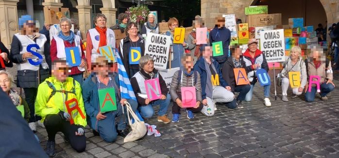 Bunt gekleidete Menschen in zwei Reihen, jeder trägt einen bunten Buchstaben. Zusammen ergeben die Buchstaben das Versprechen: "Wir sind die Brandmauer"
