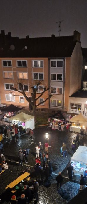 Weihnachtsmarkt bei Nacht: Blick von oben über den Weihnachtsmarkt. Man sieht nasses Pflaster, die Zelte einiger Stände und Menschen