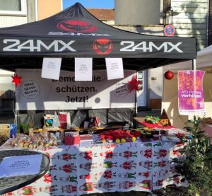 Der Stand der OMAs gegen Rechts auf dem Weihnachtsmarkt. Hinten unter dem Zelt hängt unser Banner „Demokratie schützen. Jetzt!“, vorne präsentieren wir auf weihnachtlichem Stoff unser Angebot.