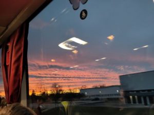 Blick aus dem Reisebus auf einen wolkigen Himmel mit schönem Abendrot. In der Scheibe spiegeln sich die Lichter aus dem Inneren des Busses.