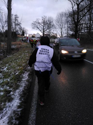 Im Morgengrauen gehen Menschen auf einer Landstraße. Letzte in der Reihe ist eine Person mit einer Weste der "OMAS gegen Rechts". Ein Auto kommt ihr mit wenig Abstand entgegen. Im Hintergrund sieht man weitere Personen, die die Landstraße entlang auf ein Dorf zulaufen.