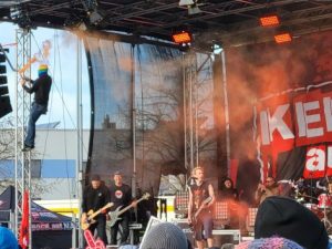 Großaufnahme des ZSK-Konzerts: Die Band steht auf der Bühne, am linken Pfeiler ist eine Person mit einem orangen Bengalo in der Hand hochgeklettert. Oranger Rauch weht an der Bühne entlang.