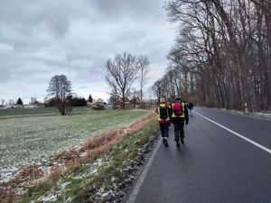 Eine Landstraße. Rechts davon steht Wald, links ein Feld, auf dem etwas Schnee liegt. Die Landstraße führt einen Hügel hoch, auf dem in der Ferne ein Dorf zu sehen ist. Auf der Landstraße laufen einige Menschen Richtung Dorf.