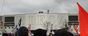 Eine große Halle, an deren Wand in großen Buchstaben RIESA steht. Auf dem Dach der Halle stehen vier Personen.