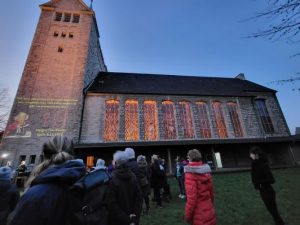 Die Martin-Luther-Kirche im Abendlicht, die hohen Fenster sind von innen erleuchtet. Ein Zitat von Margot Friedländer, Überlebende des Holocaust, ist auf den Turm projiziert. Im Vordergrund stehen Menschen auf dem Rasen.
