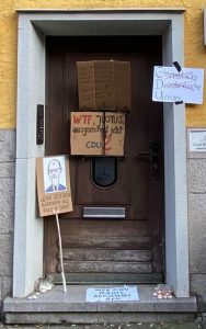 Die Eingangstür im gelben Haus der Hildesheimer CDU-Zentrale. Hier hängen mehrere Schilder. Auf einem steht „Christlich Demokratische Union", wobei „Christlich“ und „Demokratische“ durchgestrichen sind. Auf einem anderen steht „WTF, Justus, ausgerechnet jetzt CDU?“ Mit „Justus“ ist Justus Lüder gemeint, der Bundestagskandidat der CDU. Auf einem Bild ist eine Karikatur von Friedrich Merz mit seinem kleinen Rest-Haarschopf auf der Stirn, darunter steht: „Wenn sich dein Bärtchen als Frisur tarnt“. Auf dem Boden vor der Tür liegt ein Schild „Wer CDU wählt, bekommt AfD“. Ein weiteres Schild, das an der Tür hängt, ist nicht lesbar.