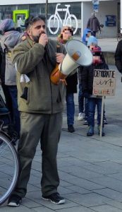 Ein Mann spricht in ein Mikrofon, das an ein Megafon angeschlossen ist. Im Hintergrund stehen mehrere Menschen, vor einem Kind steht ein Schild mit der Aufschrift "Demokratie ist bunt".