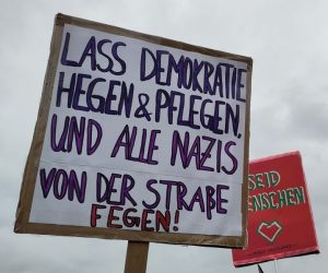 Demoplakat: "Lass Demokratie hegen & pflegen und alle Nazis von der Straße fegen!" Im Hintergrund: "Seid Menschen" als Zitat von Margot Friedländer
