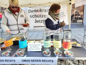 Der Infostand der Hildesheimer OMAs gegen Rechts mit einer Meinungsumfrage: Neben einem Schild „Was ist Ihnen bei der Wahl am 23. Febr. wichtig?“ stehen fünf hohe Gläser, die mit bunten Zetteln mit unterschiedlichen Aufschriften beklebt sind: „Demokratie und Rechtsstaatlichkeit“, „Klima und Umweltschutz“, „Meinungs- und Pressefreiheit“, „Soziale Gerechtigkeit und Solidarität“ sowie „Menschrenrechte und Asylrecht“. Im Hintergrund stehen zwei OMAs.