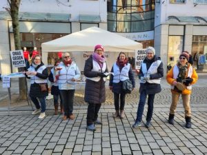 Vor dem Infostand der OMAs gegen Rechts in der Hildesheimer Fußgängerzone stehen sieben lächelnde OMAs mit weißen Westen. Einige von ihnen haben Teller mit Keksen in der Hand.