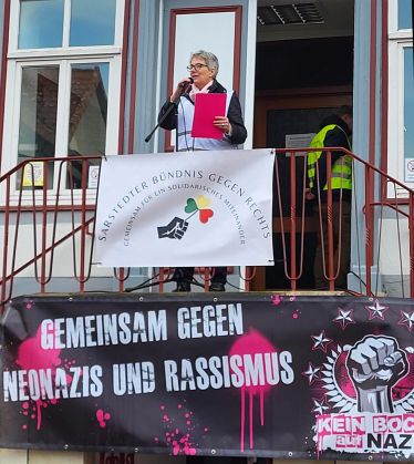 Auf dem erhöhten Treppenabsatz vor dem Sarstedter Rathaus steht eine Frau mit Mikrofon und Redemanuskript. Am Geländer hängt ein Banner mit der Aufschrift „Sarstedter Bündnis gegen Rechts“. Darunter ein schwarzes Banner, auf dem steht „Gemeinsam gegen Neonazis und Rassismus“