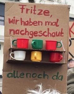 Demoplakat: "Fritze, wir haben mal nachgeschaut. Alle noch da." Am Plakat angebracht ist ein kleines Regal, auf dem sechs bunte Tassen angebracht sind.