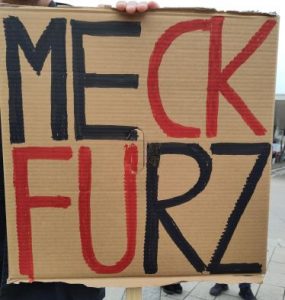 Demoplakat. Erste Zeile: MECK. Zweite Zeile: FURZ. Dabei sind ME und RZ schwarz, FU und CK rot. Liest man die passenden Farben zusammen, ergibt das FUCK MERZ.