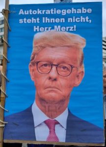 Demoplakat. Auf blauem Hintergrund ist eine Fotomontage von Donald Trump (man erkennt das an der luftigen blonden Frisur) und dem Gesicht von Friedrich Merz, mit herabgezogenen Mundwinkeln. Darüber steht: "Autokratiegehabe steht Ihnen nicht, Herr Merz!"