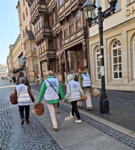 Am Hildesheimer Marktplatz. Man sieht eine Gruppe OMAs von hinten, man erkennt sie an den weißen Westen. Die Frauen gehen vor den schönen Häusern der Sparkasse entlang und tragen Körbe in den Händen.