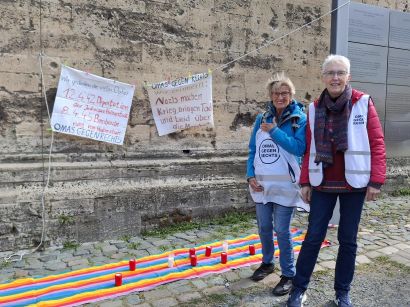 Zwei Omas gegen Rechts vor einer Kirchenwand, vor der Gedenktransparente aufgehängt sind. Auf dem Boden liegt ein langer, bunter Stoffläufer, darauf stehen einige Kerzen.