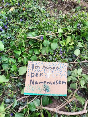 Auf dem mit grünen Efeuranken und violett blühendem Immergrün geschmückten Erdboden liegt ein kleines Pappschild mit der schön gestalteten Aufschrift "Im Namen der Namenlosen".