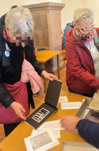 Auf einem Tisch sind Karten und Dokumente ausgebreitet. Eine Frau hält ein kleines altes Fotoalbum mit schwarzen Seiten. Sie und eine andere Frau betrachten das Foto auf der aufgeschlagenen Seite.