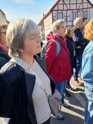 Mehrere Menschen stehen unter blauem Himmel im Freien und lauschen konzentriert einem Vortrag. Im Hintergrund ein Fachwerkhaus.