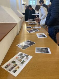 In der Gedenkstätte. An der Wand entlang steht eine Reihe Tische, darauf liegen Fotos und Dokumente von ehemaligen KZ-Insassen. Entlang den Tischen stehen Menschen und betrachten alles interessiert.