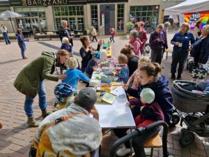An einem langen Tisch sitzen Eltern und Kinder und basteln gemeinsam. Im Hintergrund stehen weitere Menschen, die das Fest besuchen.