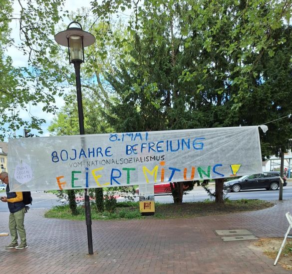 Über den Platz ist ein Banner gespannt. Aufschrift: "8. Mai. 80 Jahre Befreiung vom Nationalsozialismus. Feiert mit uns!"