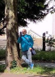 Ein Man in türkiser Jacke lehnt an einem Baum und hält filmend ein Handy in die Höhe.