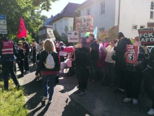 Die Kundgebung auf dem Gehweg gegenüber vom Veranstaltungshaus. Mehrere Menschen stehen herum, viele halten Schilder.