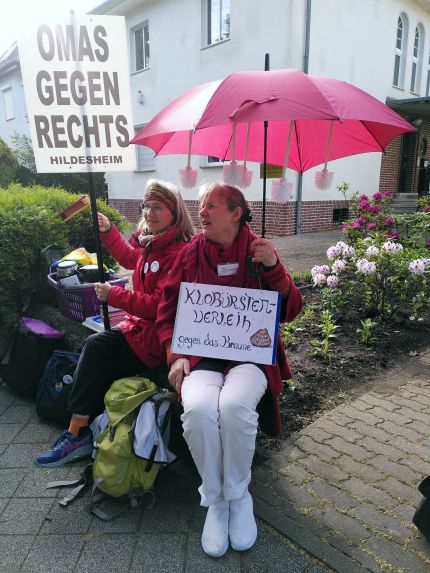 Zwei Hildesheimer OMAS GEGEN RECHTS. Die eine hält ein Schild mit der Aufschrift "OMAS GEGEN RECHTS Hildesheim", die andere trägt einen roten Schirm, von dem Klobürsten baumeln, dazu hat sie ein Schild "Klobürstenverleih gegen das Braune", daneben noch ein Häufchen.