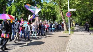 Der Demozug auf der Straße. Auf dem Gehweg daneben gehen Polizisten.