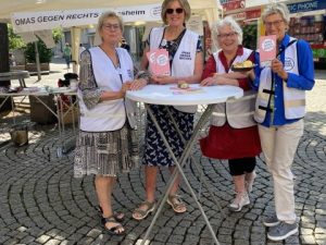 Vier Omas vor dem Stand der OMAS GEGEN RECHTS auf der 2. Meile der Demokratie in Hildesheim. Sie tragen weiße OMA-Westen, stehen vor einem Stehtisch und halten Kekse und Flyer in die Höhe.
