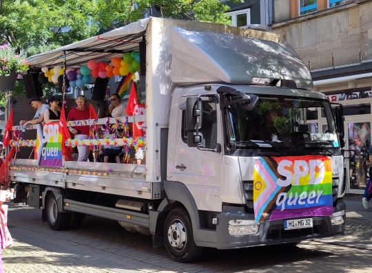 Ein Lastwagen, der an der Seite offen ist. Er ist mit regenbogenbunten Luftballons und Girlanden geschmückt. An der Seite und vorne am Kühler ist die Regenbogen-Trans-Flagge mit der Aufschrift "SPD queer" befestigt.