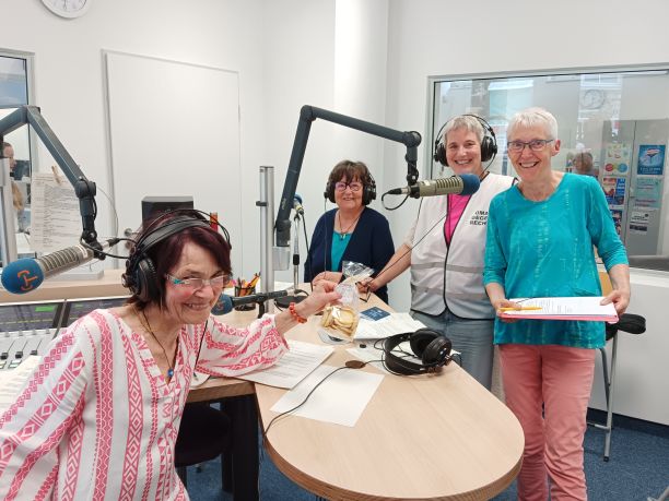 Im Radio-Studio. Links vom Tisch die Moderatorin, rechts vom Tisch drei OMAS GEGEN RECHTS, eine davon trägt OMA-Weste. Die Moderatorin und zwei OMAS tragen Kopfhörer. Die Moderatorin hält eine Tüte Kekse hoch, eine OMA hält Unterlagen in der Hand, und alle strahlen in die Kamera.