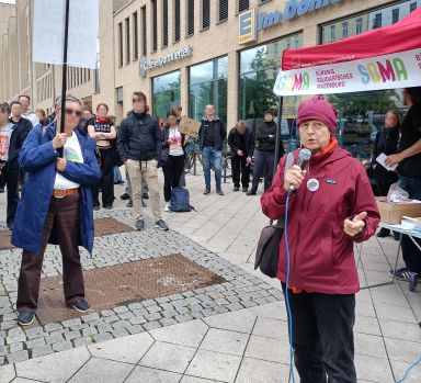 Von Zuhörern umringt steht eine Frau mit roter Jacke und roter Mütze und spricht in ein Mikrofon.