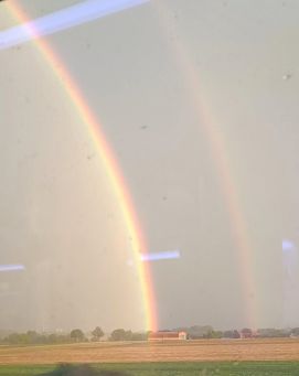 Foto aus dem fahrenden Zug: Über den Feldern ist ein leuchtender Doppel-Regenbogen zu sehen.