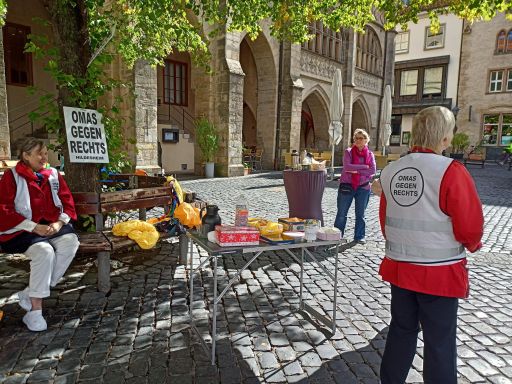 Sonnige Stimmung auf dem Hildesheimer Marktplatz. Tische mit Kaffee und Keksen stehen auf dem Kopfsteinpflaster, zwei OMAs mit Westen warten auf Besucher, im Hintergrund steht eine weitere Frau.
