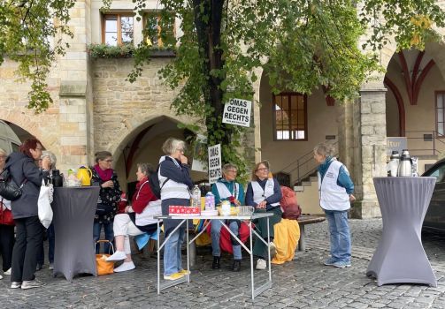 Unter der großen Linde stehen einige OMAs mit Westen, vor Baum und Bank sind Tische mit Kaffee und Keksen aufgestellt. Links im Bild findet ein Gespräch statt, einen weitere Frau trinkt etwas.