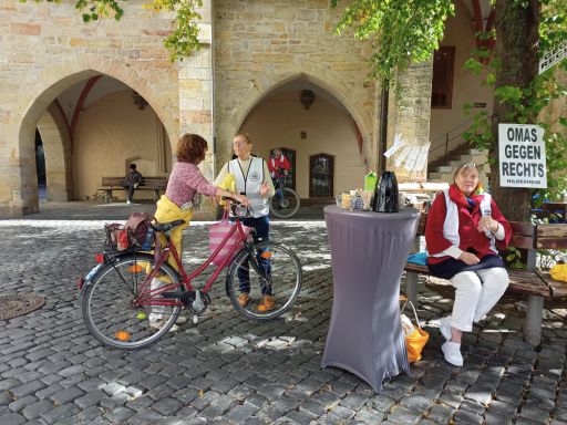 Eine OMA mit Weste sitzt auf der Bank, die um den Baum gebaut ist. Neben ihr ein Tisch mit Kaffeeangebot. Im Hintergrund spricht eine weitere OMA mit einer Frau mit Fahrrad.