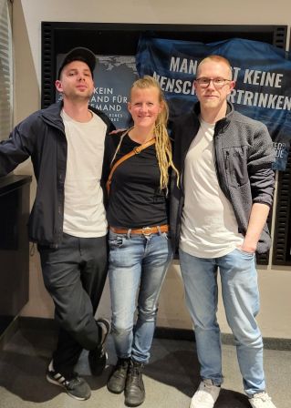 Die beiden Filmemacher und die junge Frau, die Rettungseinsätze im Mittelmeer fliegt, posieren im Kino vor dem Kinoplakat und der Fahne mit der Aufschrift "Man lässt keine Menschen ertrinken. Punkt." Der Hintergrund ist aber zum Teil von den Köpfen verdeckt.