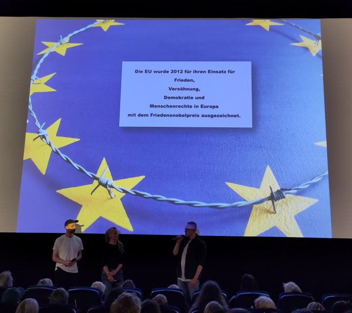Blick auf die große Kinoleinwand. Im Vordergrund Sitzreihen und Köpfe, vor der Leinwand stehen die drei Gäste, die Leinwand zeigt die blaue Europaflatte mit den gelben Sternen. Aber auf dem Kreis, den die Sterne bilden, liegt ein Stacheldraht, Auflagepunkte der Stacheln sind die Sterne. In der Mitte auf weißem Hintergrund der Hinweis: "Die EU wurde 2012 für ihren Einsatz für Frieden, Versöhnung, Demokratie und Menschenrechte in Europa mit dem Friedensnobelpreis ausgezeichnet."