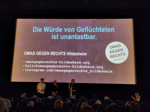 Blick auf die große Kinoleinwand. Im Vordergrund Sitzreihen und Köpfe, vor der Leinwand stehen die drei Gäste, auf der Leinwand ist eine Botschaft der OMAS GEGEN RECHTS zu lesen: "Die Würde von Geflüchteten ist unantastbar." Dazu diverse Kontaktadressen.