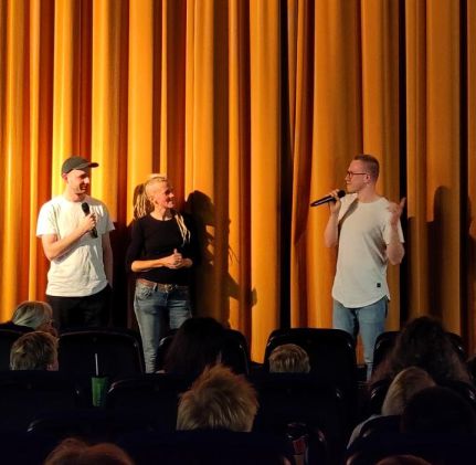 Vor einem in Falten geworfenen Kinovorhang in Goldtänen stehen drei Menschen: Links und rechts je ein junger Mann in Jeans und weißem T-Shirt. Sie halten Mikrofone in den Händen. In der Mitte eine junge Frau in Jeans und schwarzem Shirt. Im Vordergrund sieht man Sitzreihen und Köpfe von Menschen im Publikum.