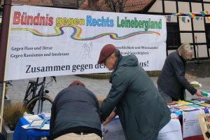 Der Stand des „Bündnis gegen Rechts Leinebergland“ in Gronau. Auf dem Schild steht „Gegen Hass und Hetze“, „Gegen Rassismus und Intoleranz“, „Menschlichkeit und Miteinander“ und (teils verdeckt) „... und Gerechtigkeit“. Im Drei Personen sind mit dem Infomaterial beschäftigt, das am Stand ausliegt.