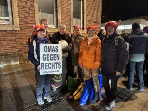 Nachts steht das Grüppchen der Hildesheimer OMAS GEGEN RECHTS mit Demoplakaten auf der Straße. Wer die Ecke kennt, weiß, dass es in der Nähe des Hildesheimer Hauptbahnhofs ist.