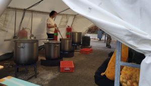 Ein großes, offenes weißes Zelt, in dem ein junger Mann im T-Shirt vor vier riesigen Kochtöpfen steht, in einem rührt er. Vorne rechts liegen Kartoffelsäcke bereit.