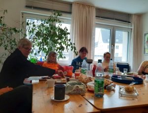 An einem Holztisch sitzen einige OMAS und wärmen sich auf. Auf dem Tisch stehen Getränke, Kuchen und Kekse.