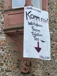 Am Erker eines historischen Gebäudes aus groben Steinen hängt ein Plakat: "#NieWiederIstJetzt Komm rein! Wir haben: Suppe, Toiletten, Tee"