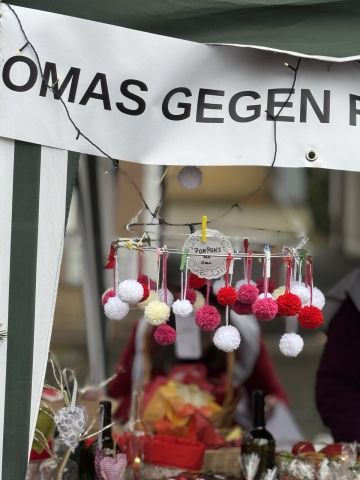 Detailaufnahme: An einem feinen Metallring hängen viele kleine Pompons in Rot- und Weißtönen. Oben schlängelt sich eine Lichterkette durchs Bild, und ganz oben sieht man einen Teil des OMAS-Banners.