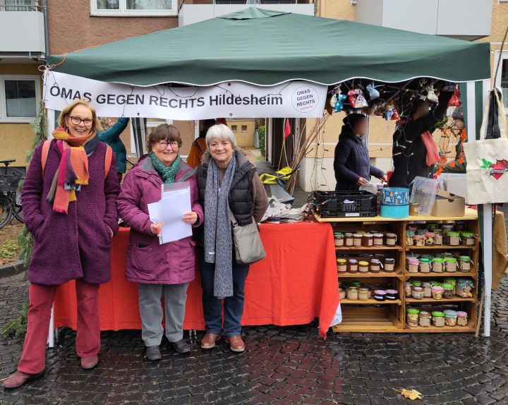 Drei Hildesheimer OMAS haben sich vor dem Stand der OMAS GEGEN RECHTS aufgestellt. Er ist unter einem grünen Pavillon, oben hängt ein Banner „OMAS GEGEN RECHTS Hildesheim“. Im rechten Teil des Pavillons ist der Stand der Eltern gegen Rechts. Sie haben ein Holzregal mit vielen Marmeladengläsern.